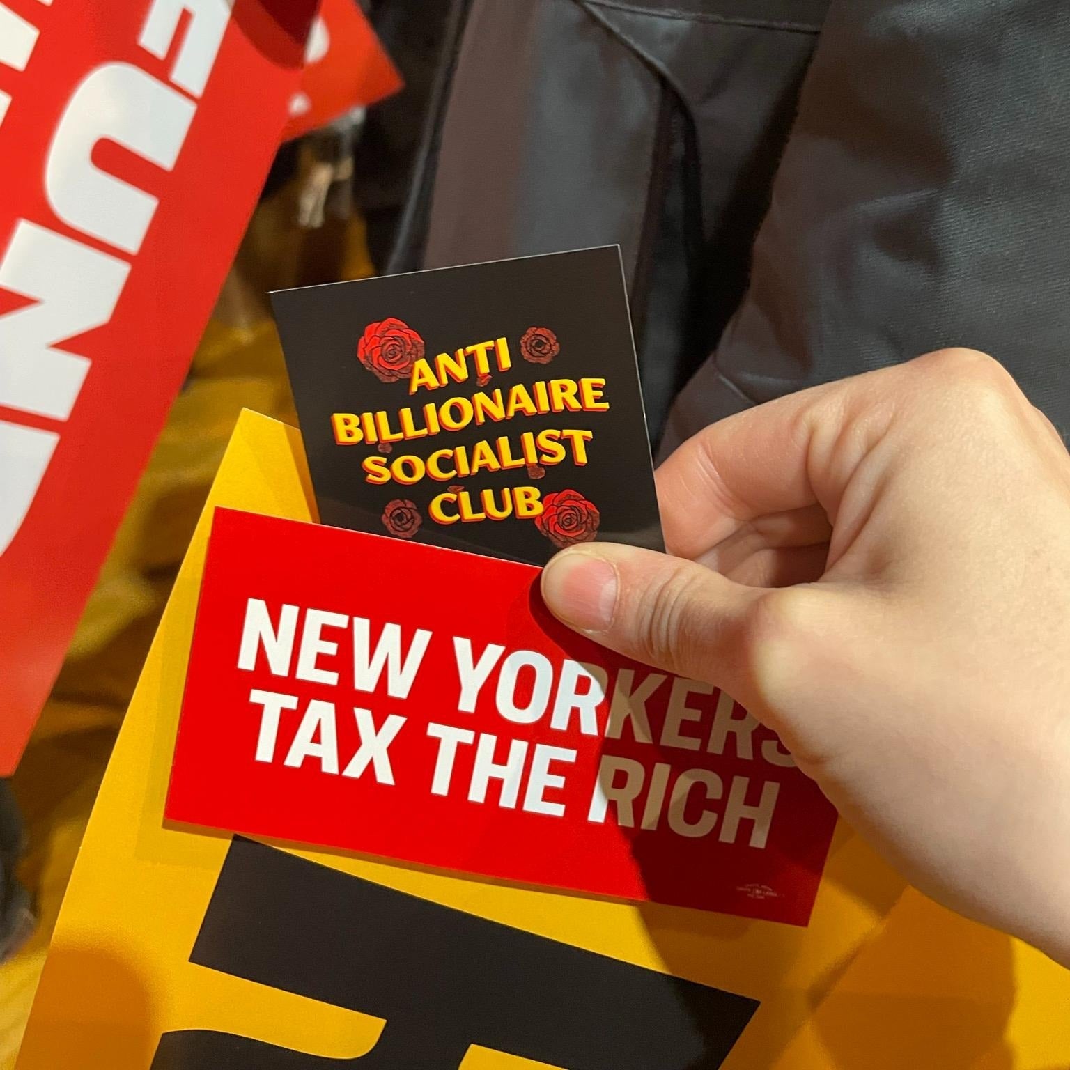 Person holding a red 'New Yorkers Tax the Rich' sticker and a 'Anti Billionaire Socialist Club' sticker.